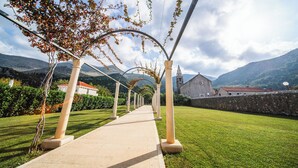 A pathway lined with columns leading to a building with a clock tower, surrounded by greenery and mountains. - Vila Riva Apartments (Dubrovacko Primorje)