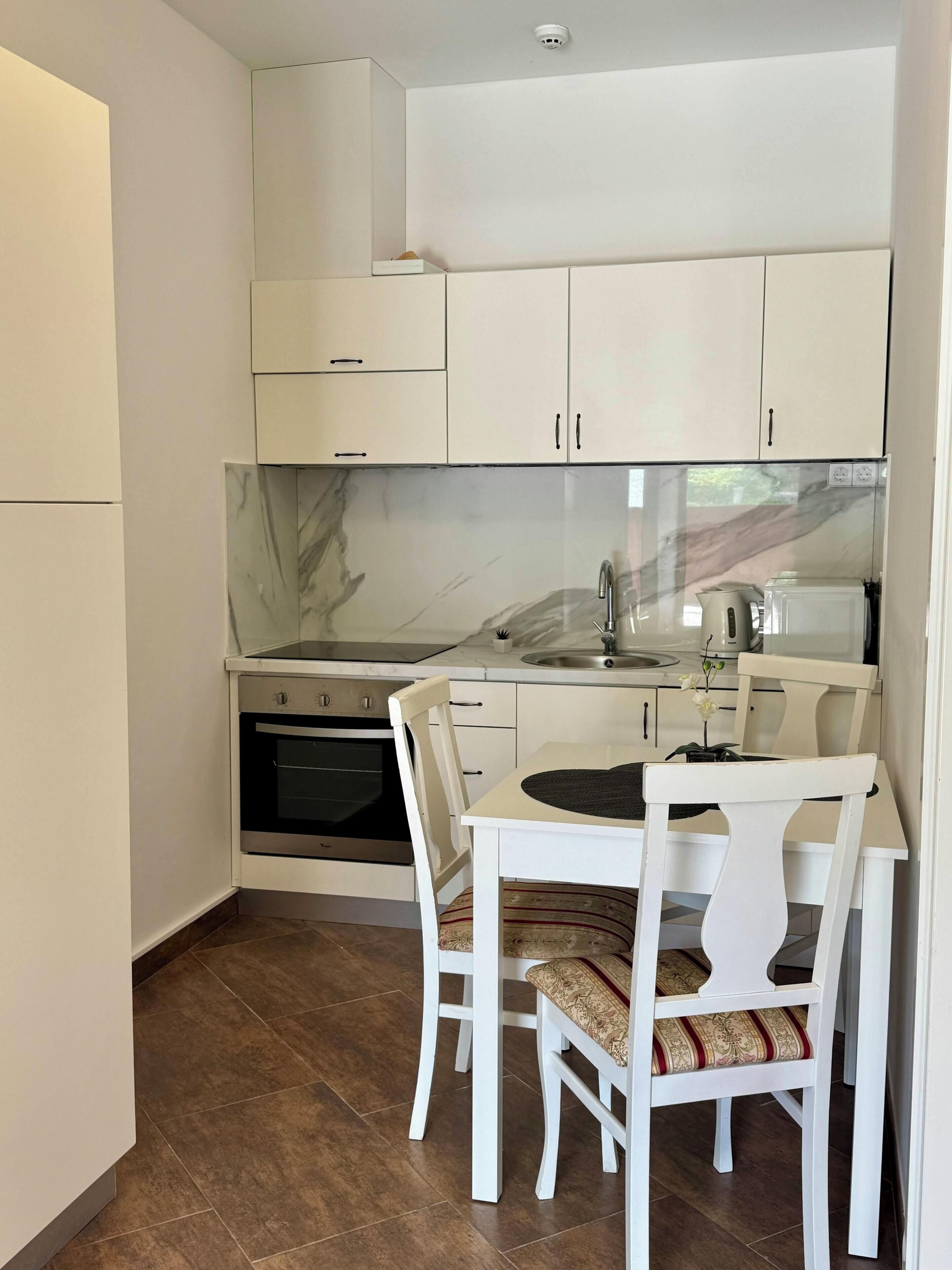 Una cucina compatta con mobili bianchi, uno schienale in marmo, forno integrato e un'area pranzo con tavolo e sedie bianche.