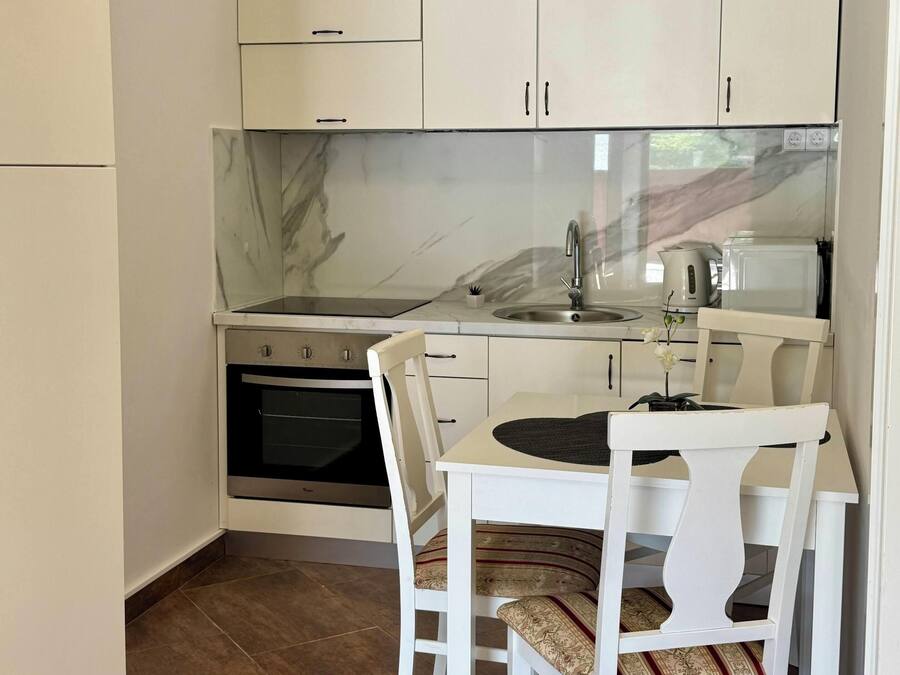 Una cocina compacta con armarios blancos, salpicadero de mármol, horno integrado y un comedor con mesa y sillas blancas.
