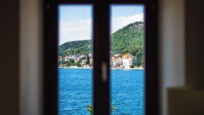 A view through a window of a coastal town with buildings and a hill in the background. - Vila Riva Apartments (Dubrovacko Primorje)