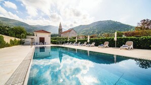 A swimming pool with lounge chairs, a small building, and a church in the background. - Vila Riva Apartments (Dubrovacko Primorje)