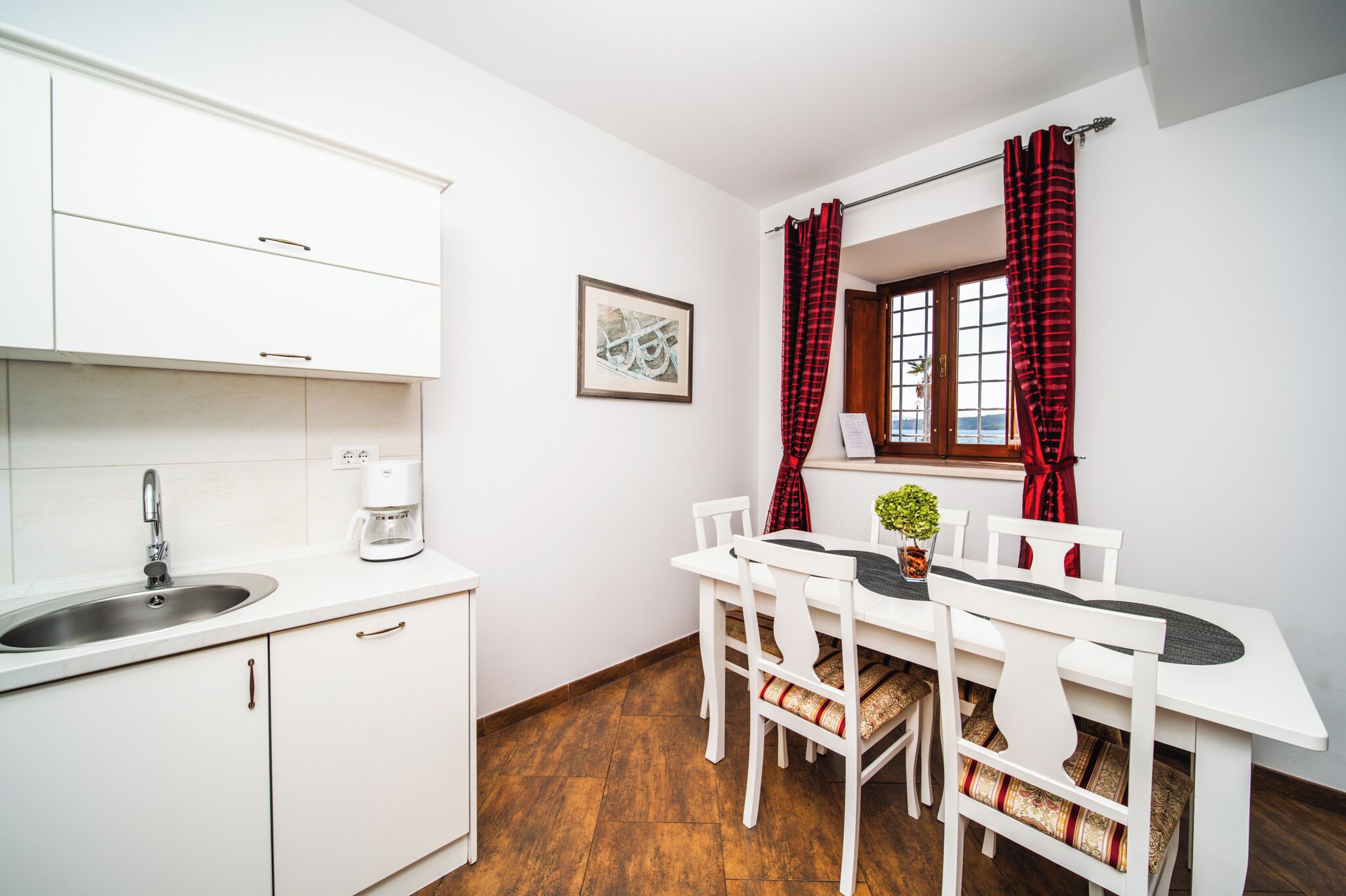 A kitchen with white cabinets, a sink, and a coffee maker. There is a dining table with white chairs and a potted plant.