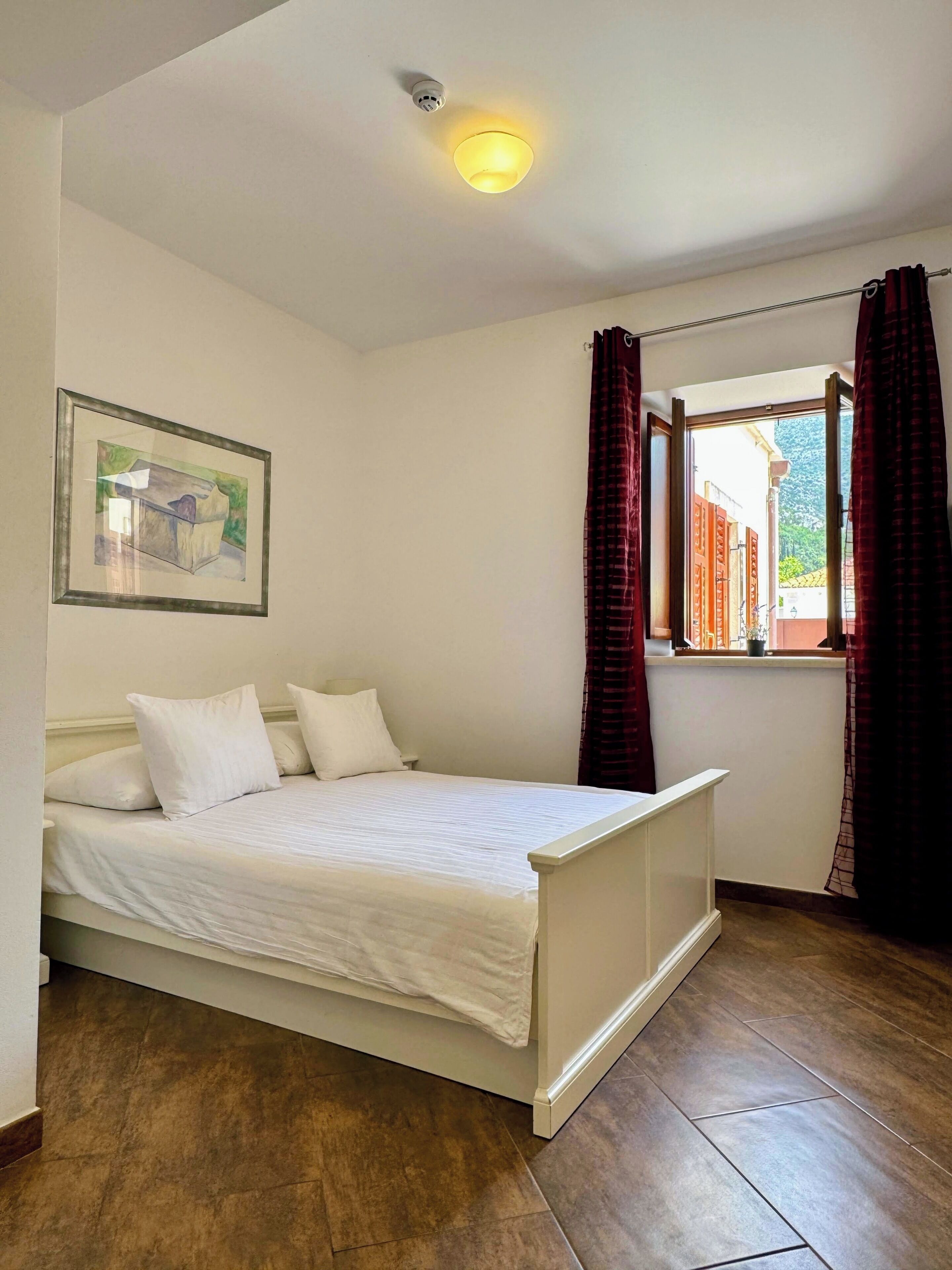 Ein Schlafzimmer mit einem Bett, einem Fenster mit Vorhängen, einer Deckenleuchte und einem gerahmten Bild an der Wand.