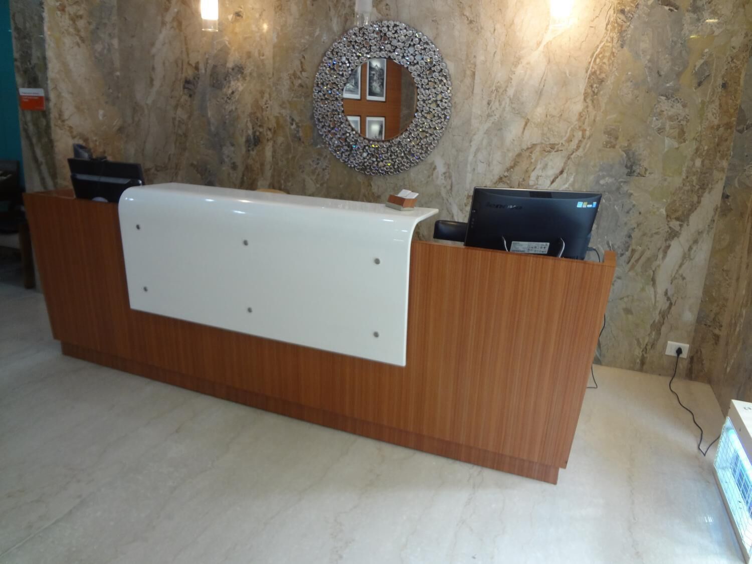 Un mostrador de recepción con una pared de mármol al fondo, un espejo decorativo y un monitor de computadora sobre el mostrador.