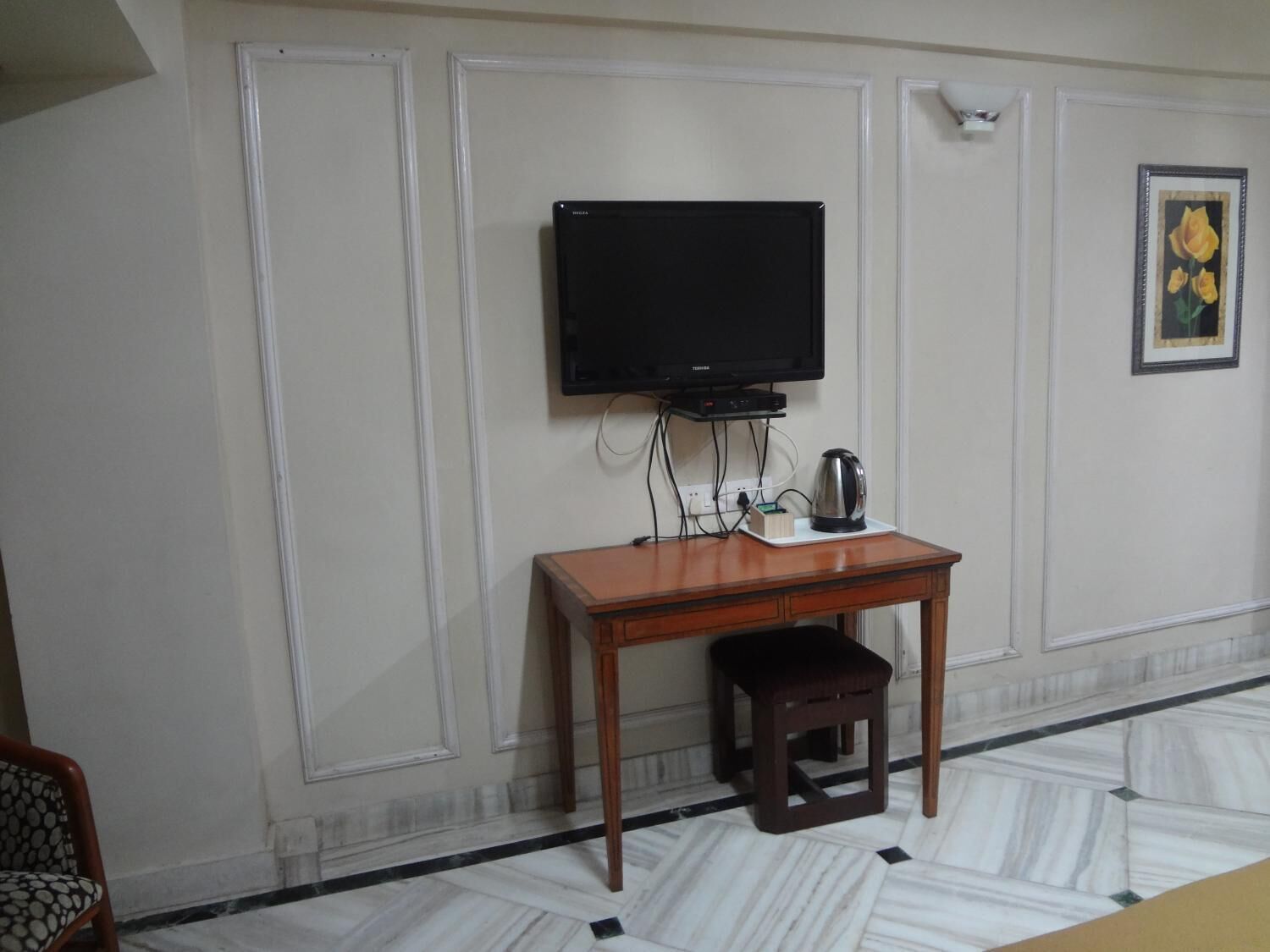 A room with a television mounted on the wall, a small wooden table with a kettle, and a framed picture on the wall.