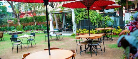 An outdoor seating area with tables and chairs, red umbrellas, and potted plants.