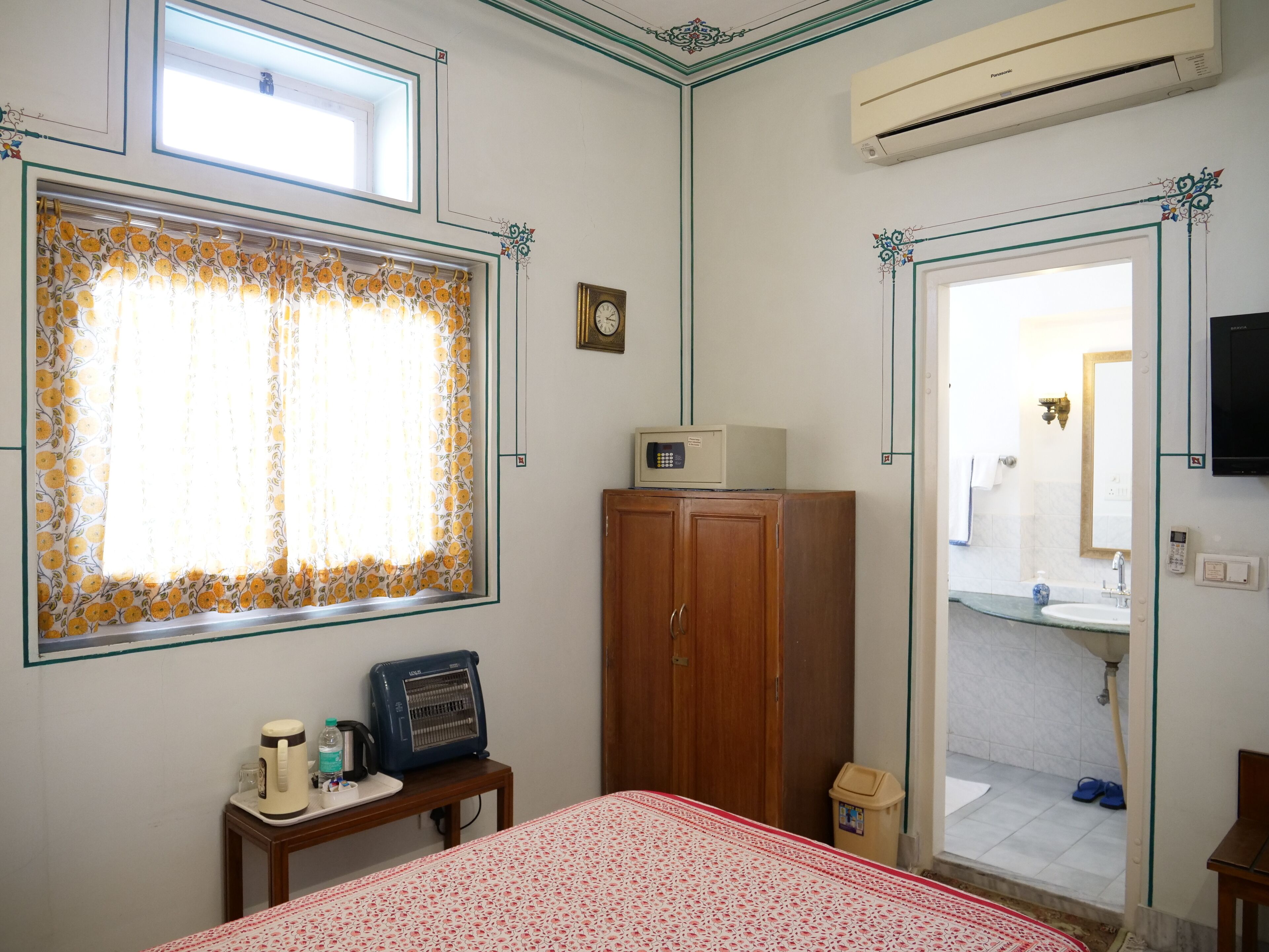 A room with a bed, a wooden wardrobe, a small table with a kettle and a radio, a window with floral curtains, and an air conditioning unit.