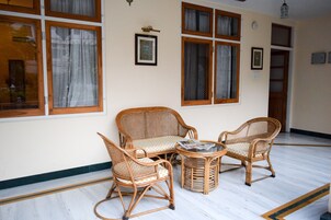 Una habitación con dos sillas de mimbre, una mesita pequeña, ventanas amplias con cortinas y una puerta de madera.