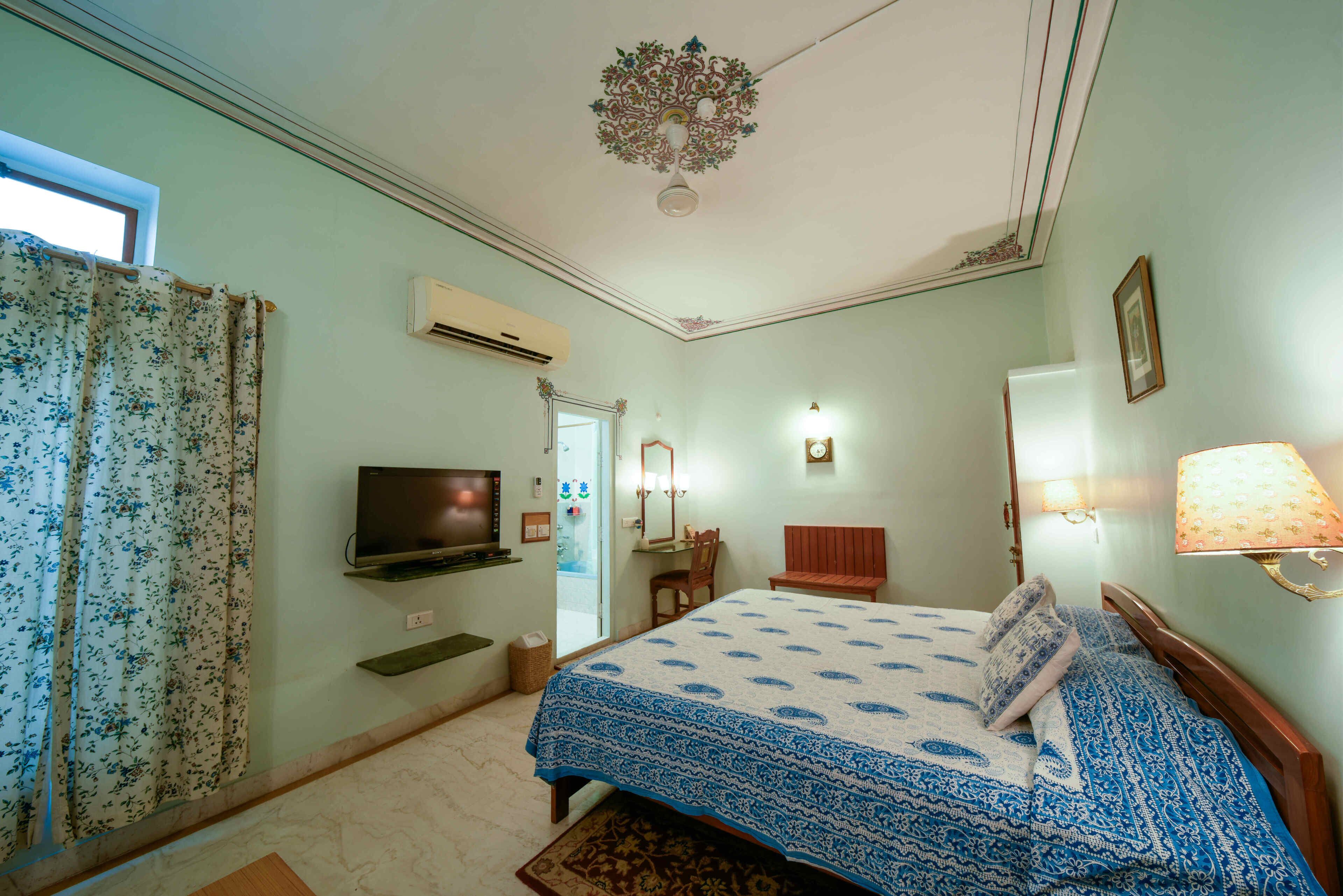 Ein Schlafzimmer mit Bett, Fernseher, Wand-Klimaanlage, Fenster mit Vorhängen, Nachttischlampe und einem kleinen Tisch.