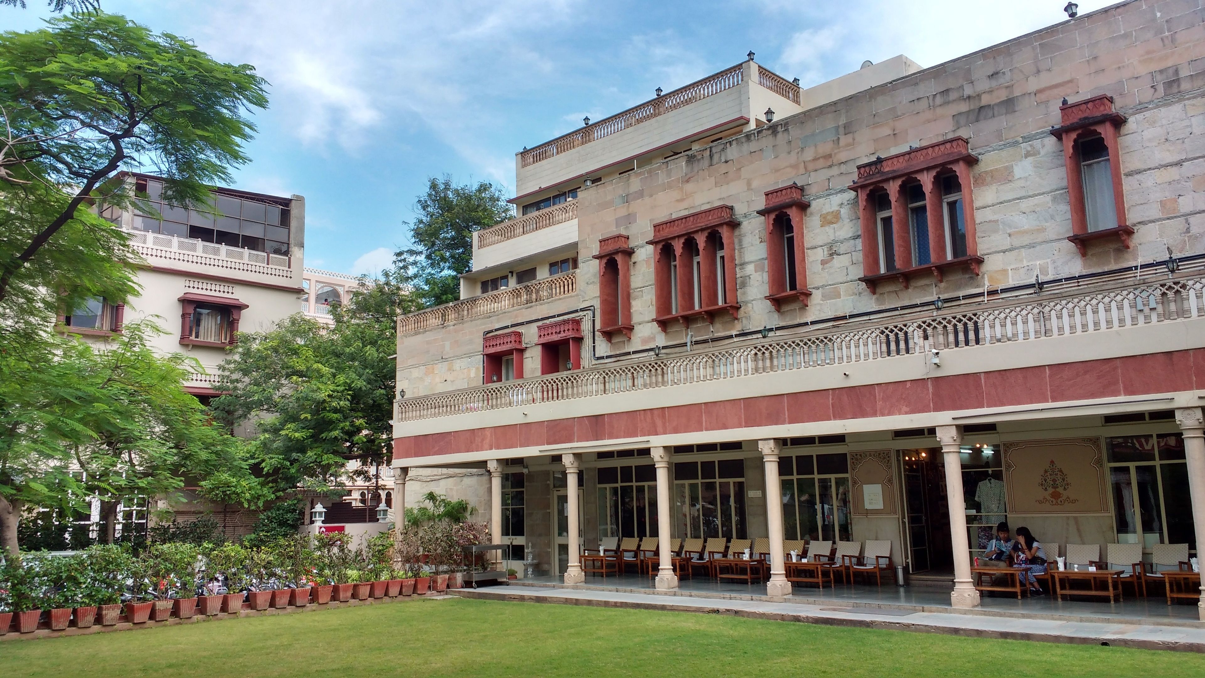 A multi-story building with a covered outdoor seating area, columns, and a garden with potted plants.