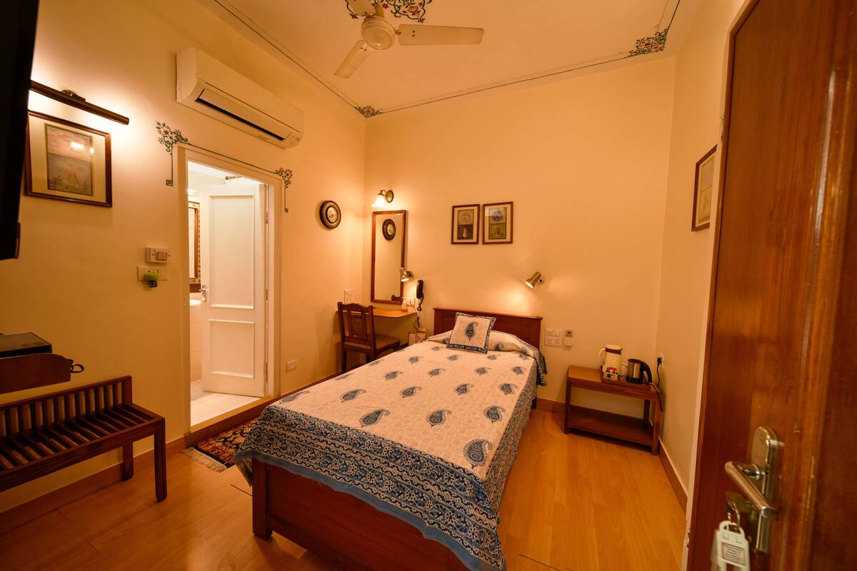Un dormitorio con cama, mesita de noche, banco y una puerta que da a otra habitación.