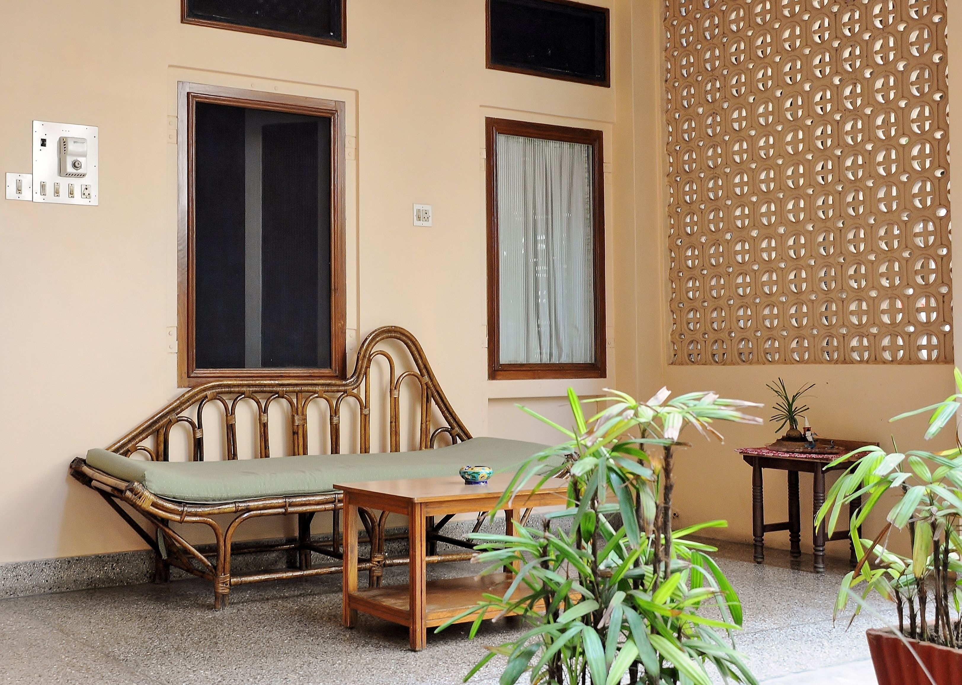 A room with a rattan sofa, a wooden table, two windows with curtains, a decorative wall panel, and a potted plant.