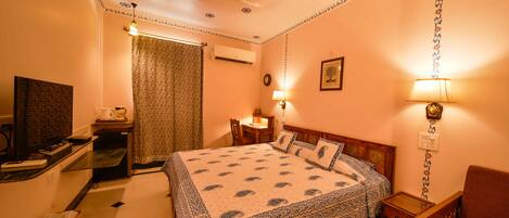Quarto de hotel com cama, escrivaninha, cadeira, abajur e televisão.