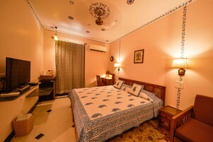 Quarto de hotel com cama, escrivaninha, cadeira, abajur e televisão.