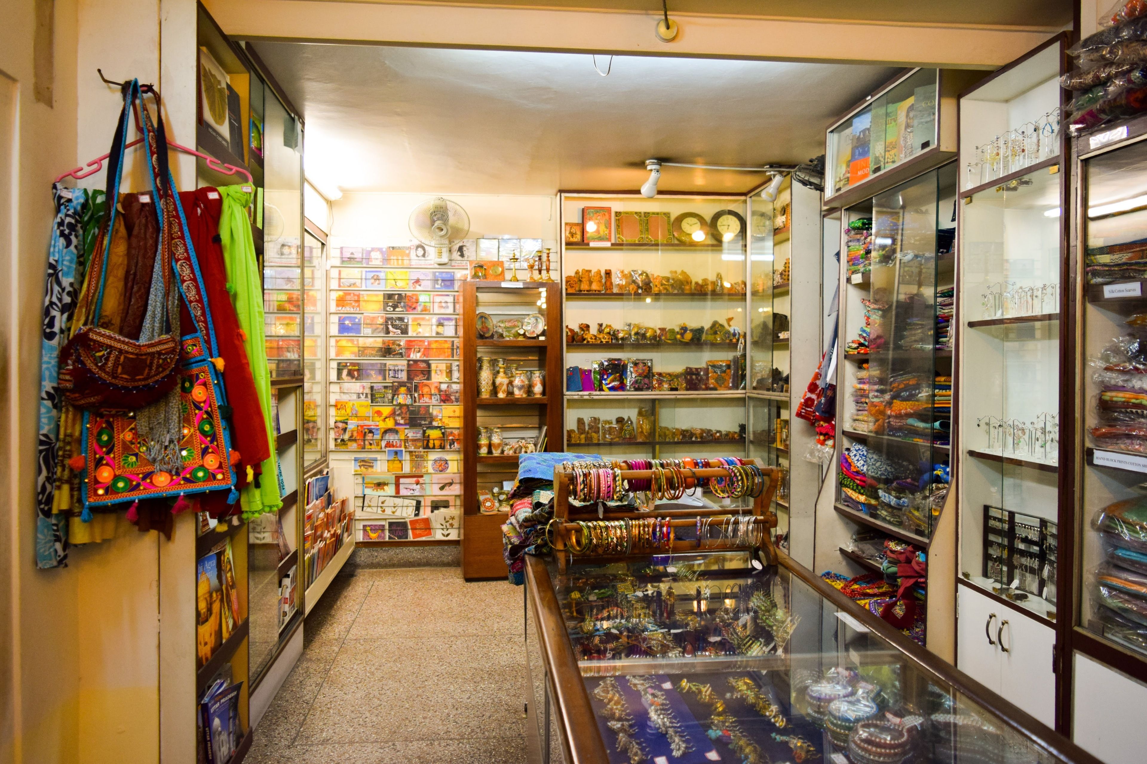 A narrow, well-lit shop with shelves stocked with various items including textiles, jewelry, and decorative objects.