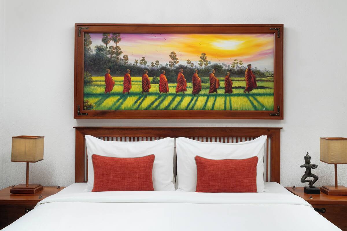 A hotel room with a bed, two nightstands, a painting of monks, and a statue.