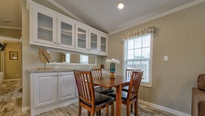 A dining area with a wooden table and chairs, white cabinets with glass doors, and a window with blinds. - Club Naples RV Resort (Naples)