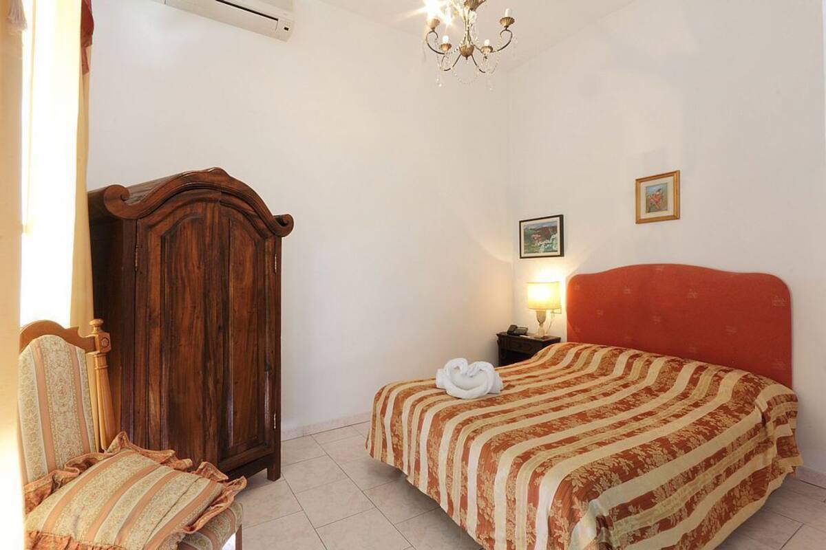 A bedroom with a wooden wardrobe, a bed with a red headboard, a chair, and a nightstand with a lamp.