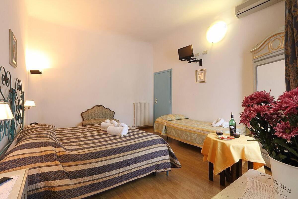 A hotel room with two beds, a small table with a bottle and glasses, a vase of flowers, and a wall-mounted TV.
