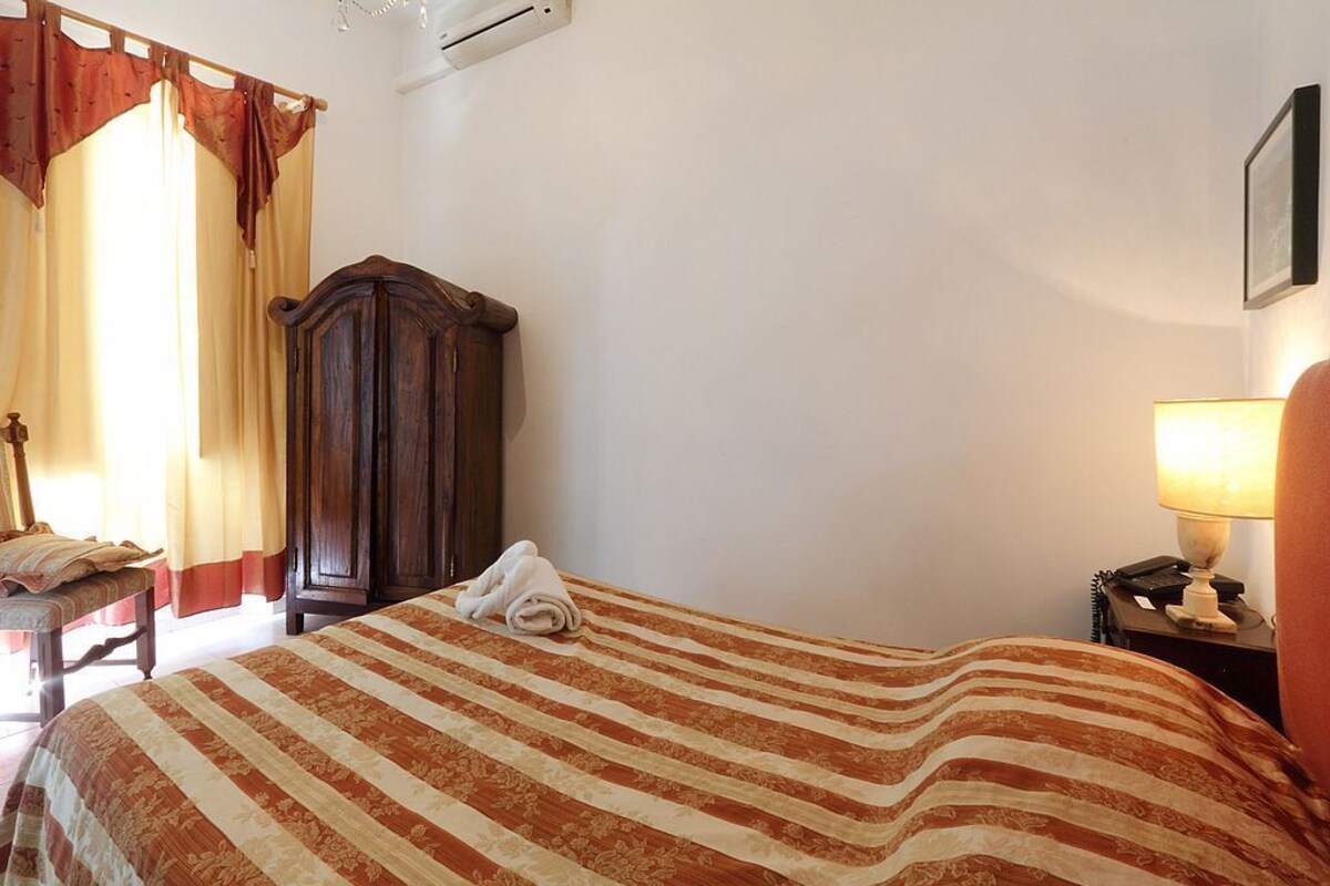 A hotel room with a bed, a wooden wardrobe, a desk with a lamp, and a chair.
