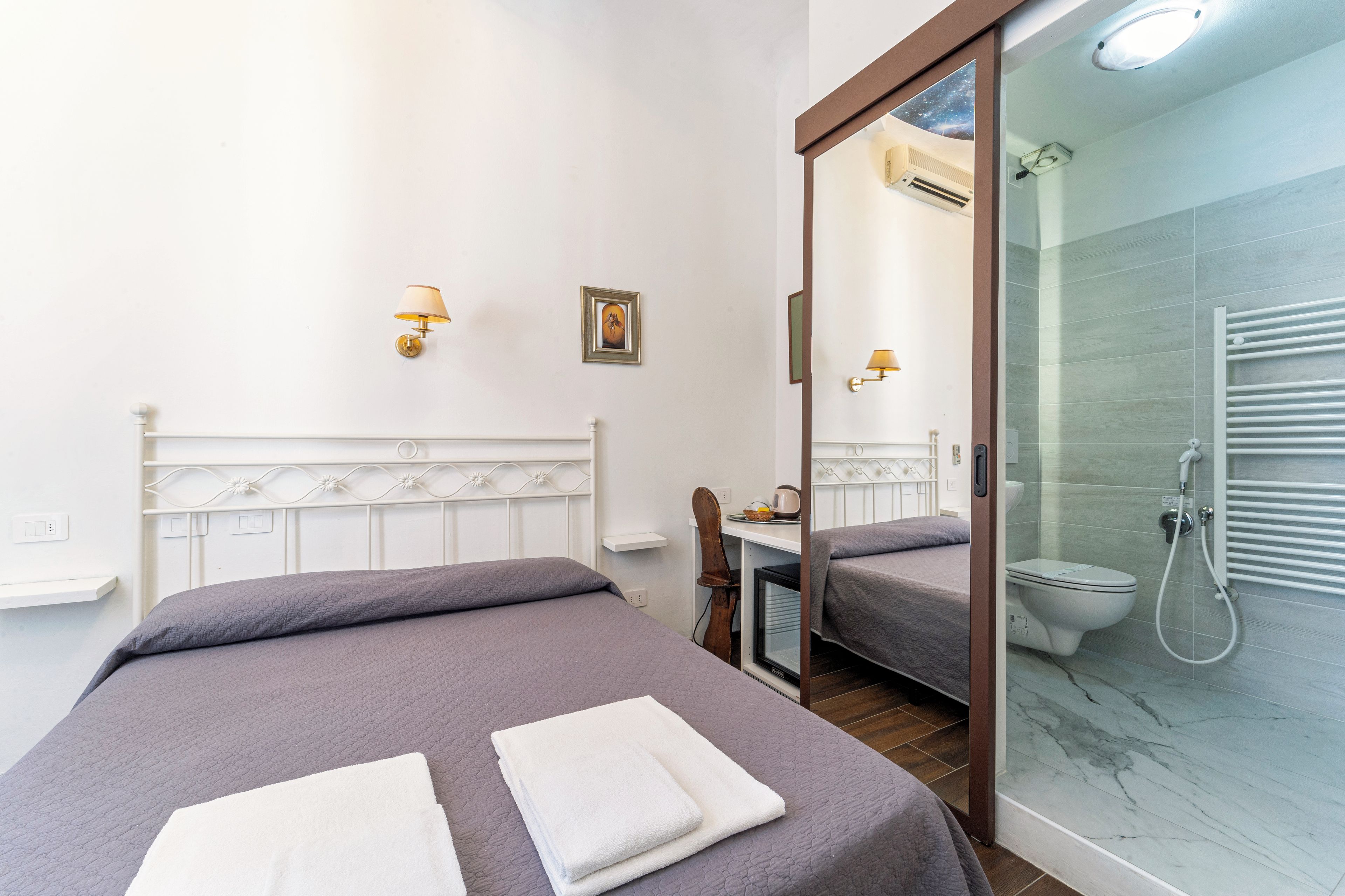 A hotel room with a bed, a bathroom with a shower, and a mirror reflecting the room.