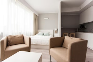 A modern hotel room with a bed, two armchairs, a small table, a kitchenette, and a large window with curtains.