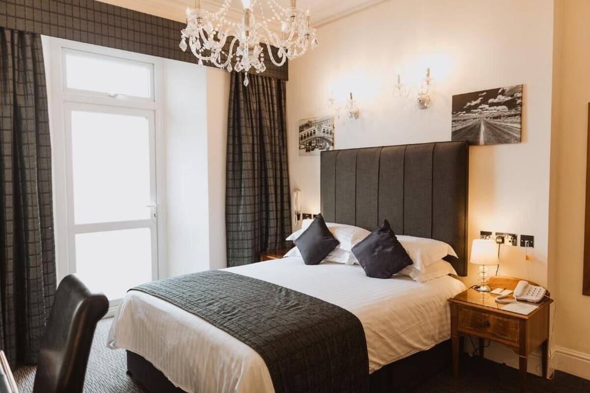 A hotel room with a large bed, a chandelier, a nightstand with a phone, and a window with curtains.