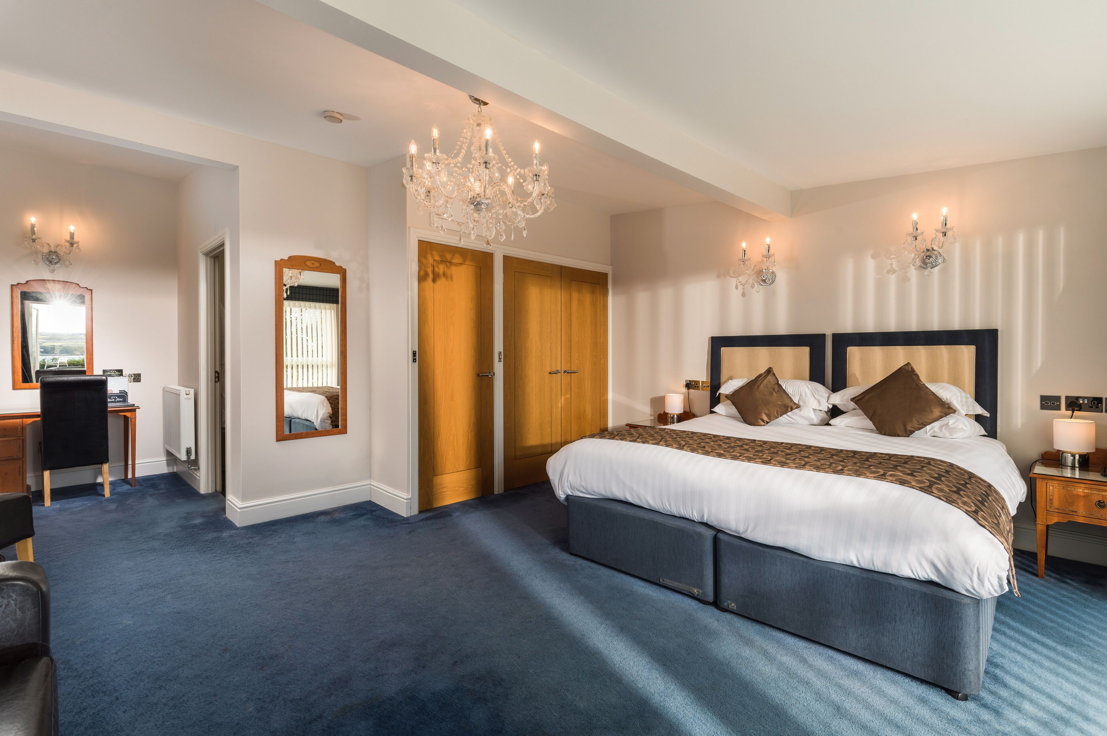 A hotel room with a large bed, a desk, a chair, a chandelier, and a wardrobe.