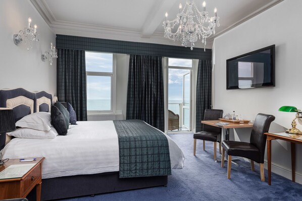 Quarto de hotel com uma cama grande, uma escrivaninha com cadeiras, um candelabro e uma televisão na parede.