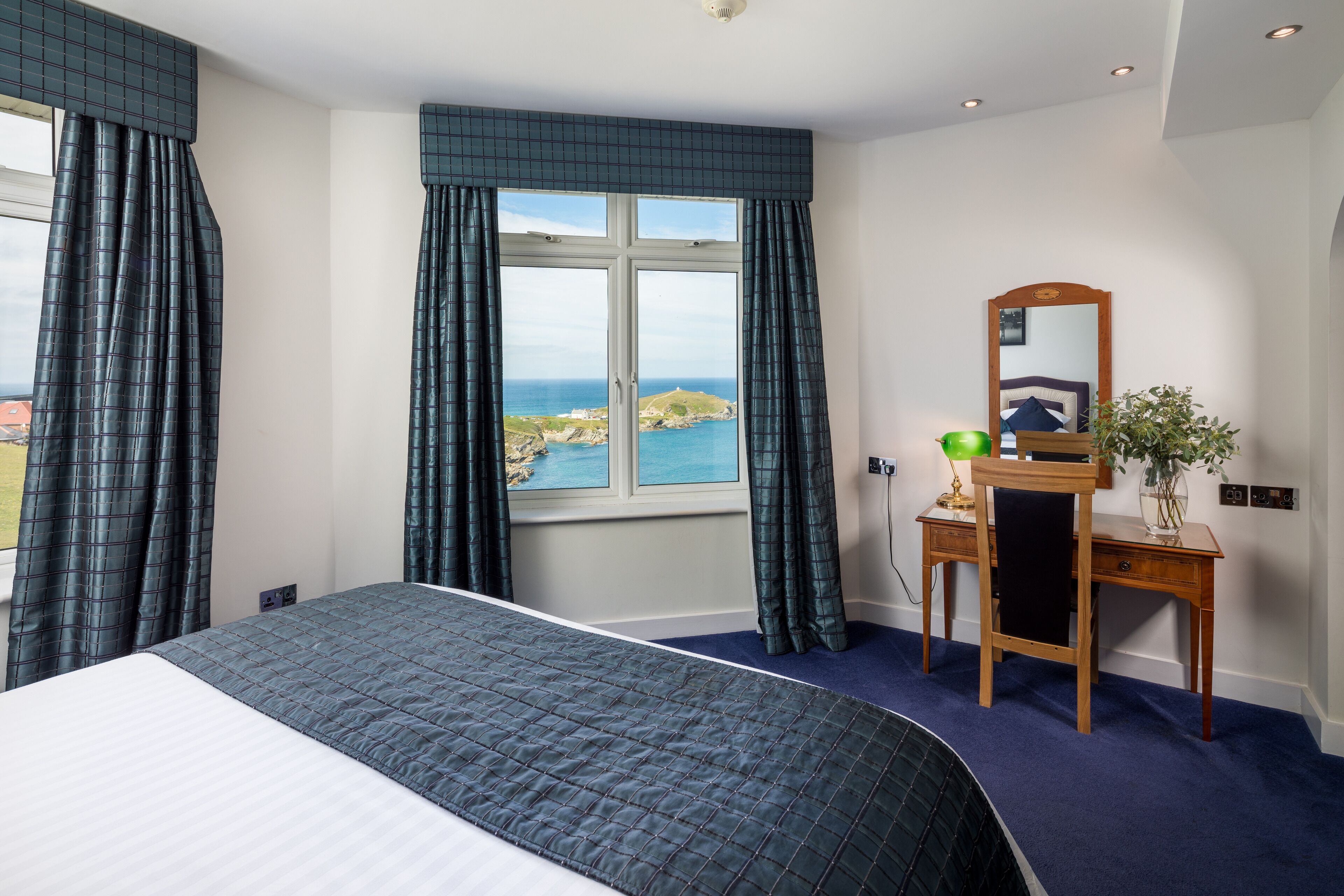 A bedroom with a large bed, a desk with a mirror, and a view of the sea.