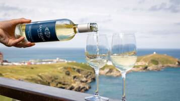 A hand pouring wine into two glasses on a balcony overlooking a coastal landscape.