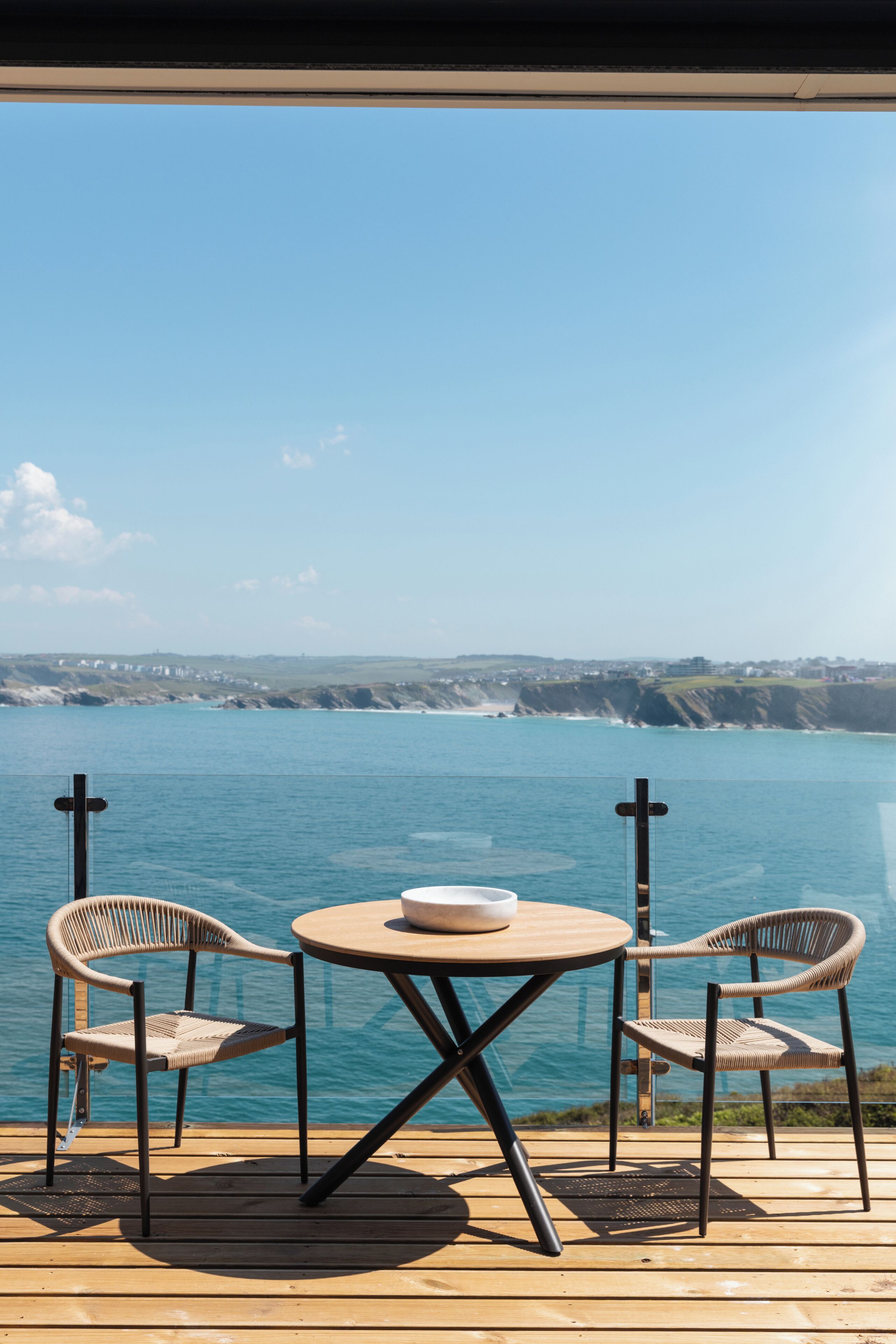 Eine Holzterrasse mit einem runden Tisch und zwei Korbstühlen, mit Blick auf ein Gewässer und eine entfernte Küstenlinie.