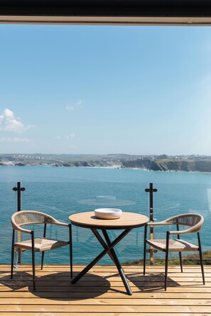 A wooden deck with a round table and two wicker chairs, overlooking a body of water and a distant coastline. - The Atlantic Hotel (Newquay)