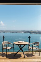 Una terraza de madera con una mesa redonda y dos sillas de mimbre, con vista a una masa de agua y una costa distante.
