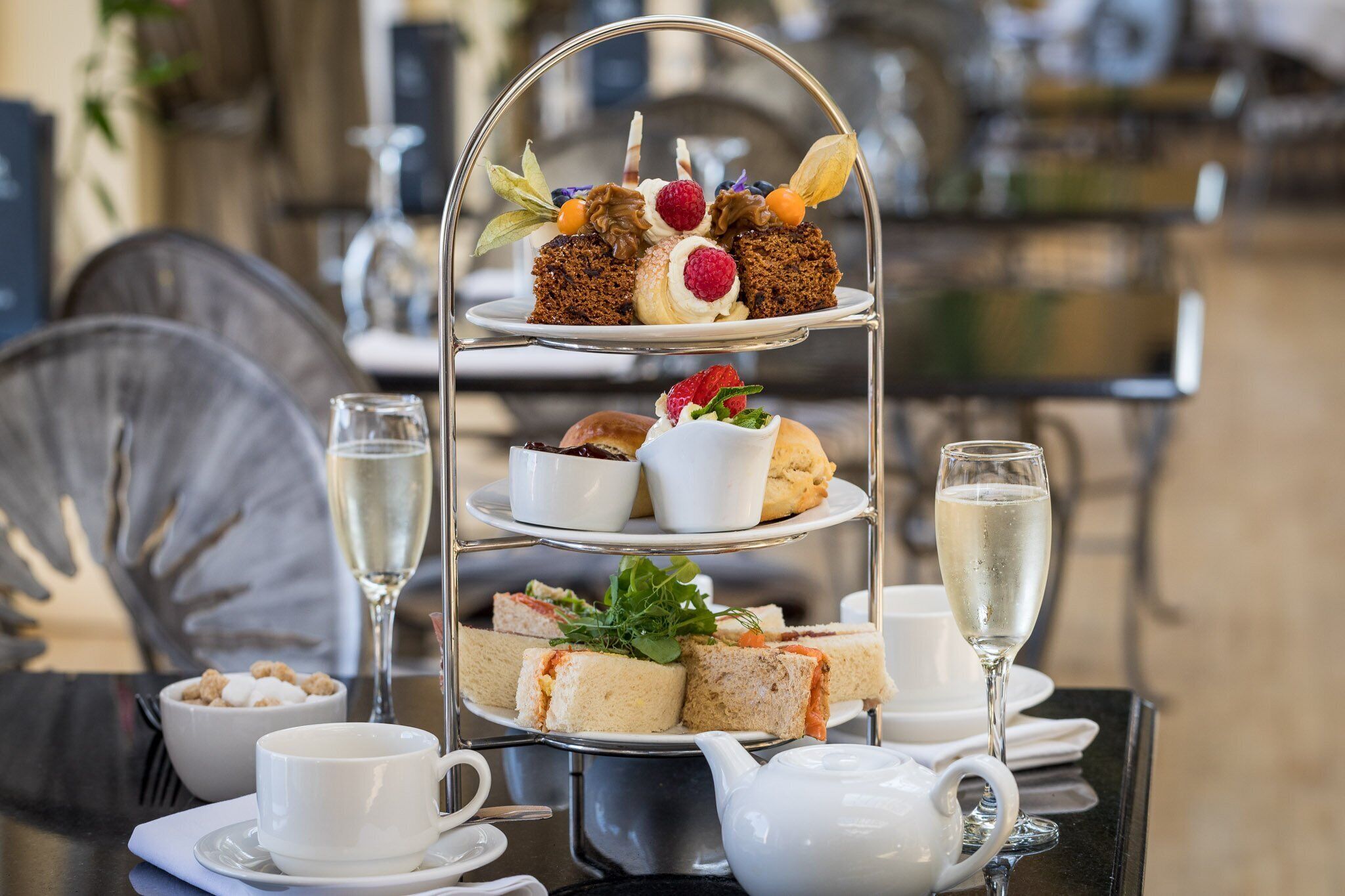A three-tiered dessert stand with various pastries and sandwiches, accompanied by white cups and a teapot on a table.