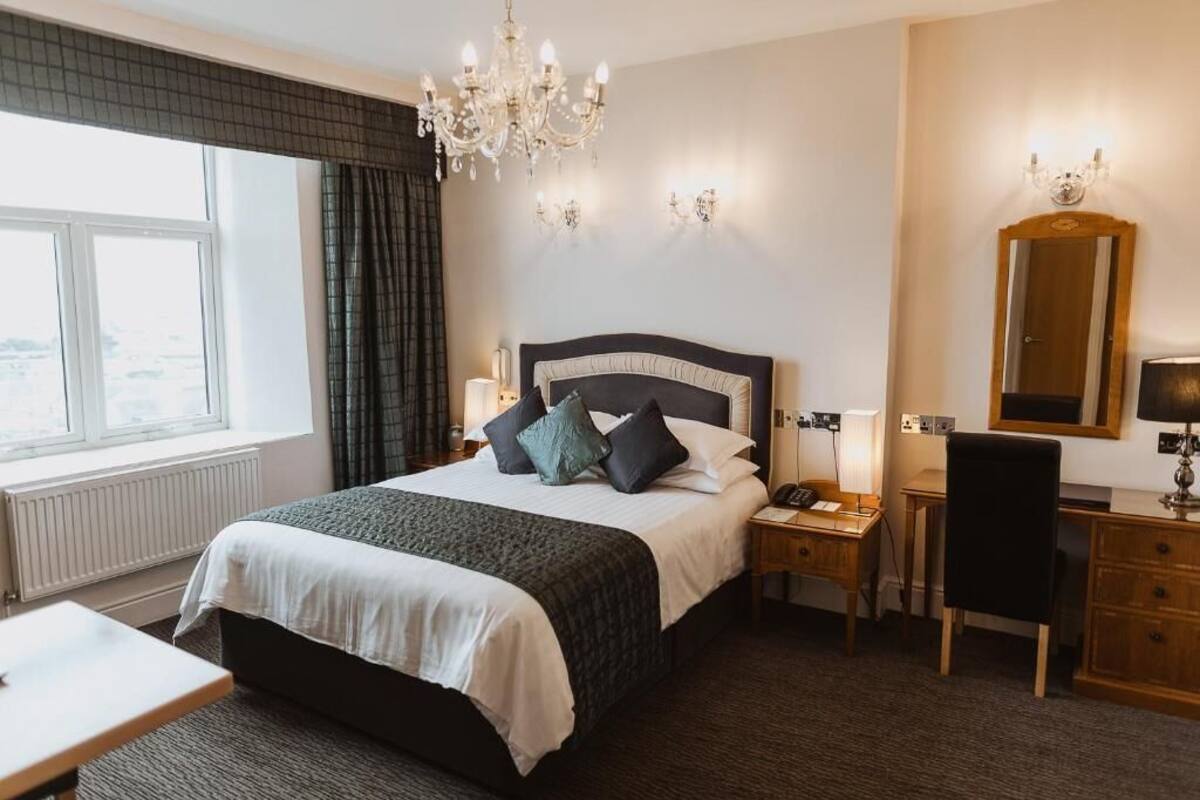 A hotel room with a large bed, a desk, a chair, a desk lamp, a telephone, a mirror, and a chandelier.