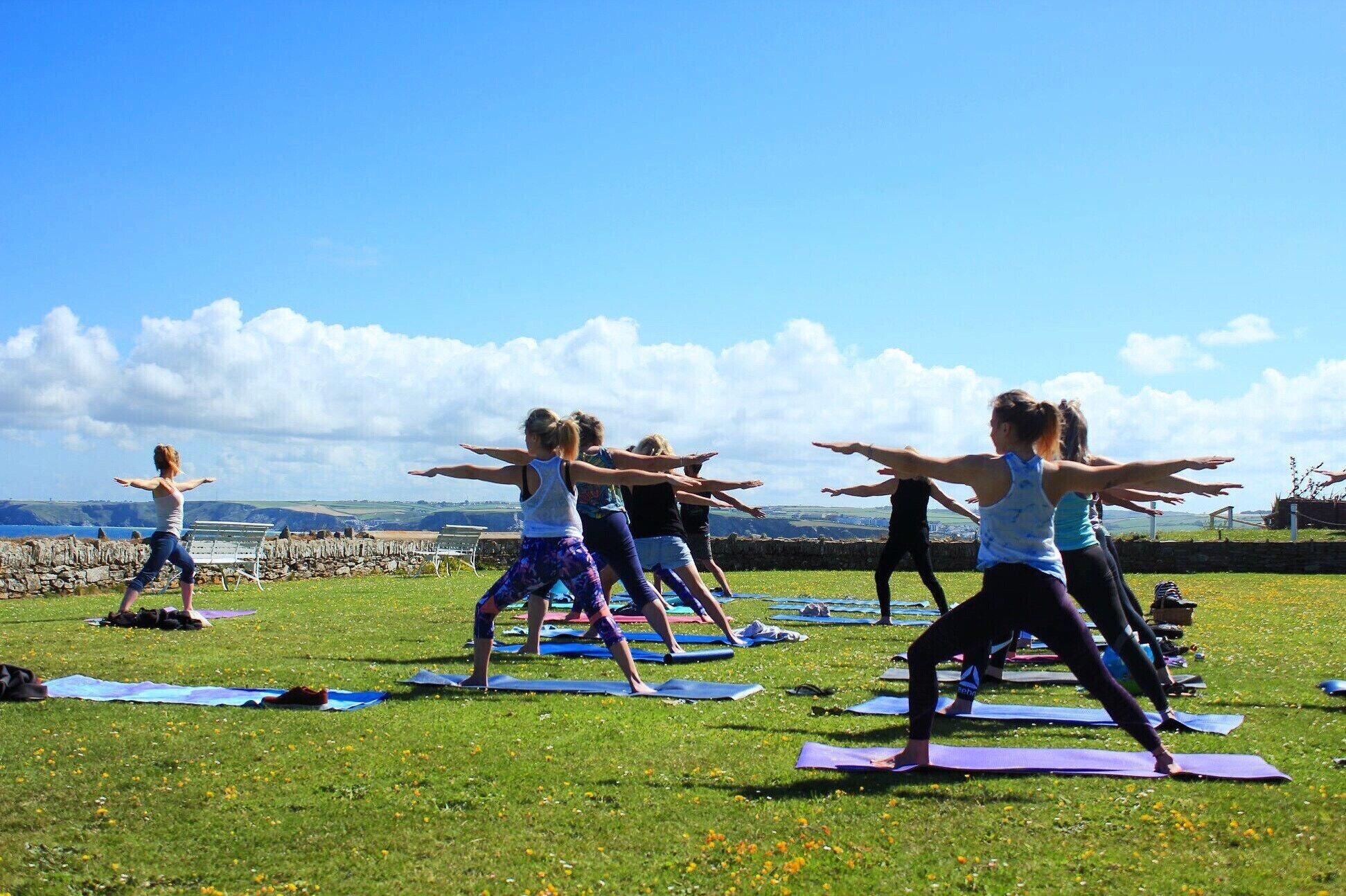 A group of people performing yoga on mats in an open grassy area with a scenic view of hills and a bridge in the background.