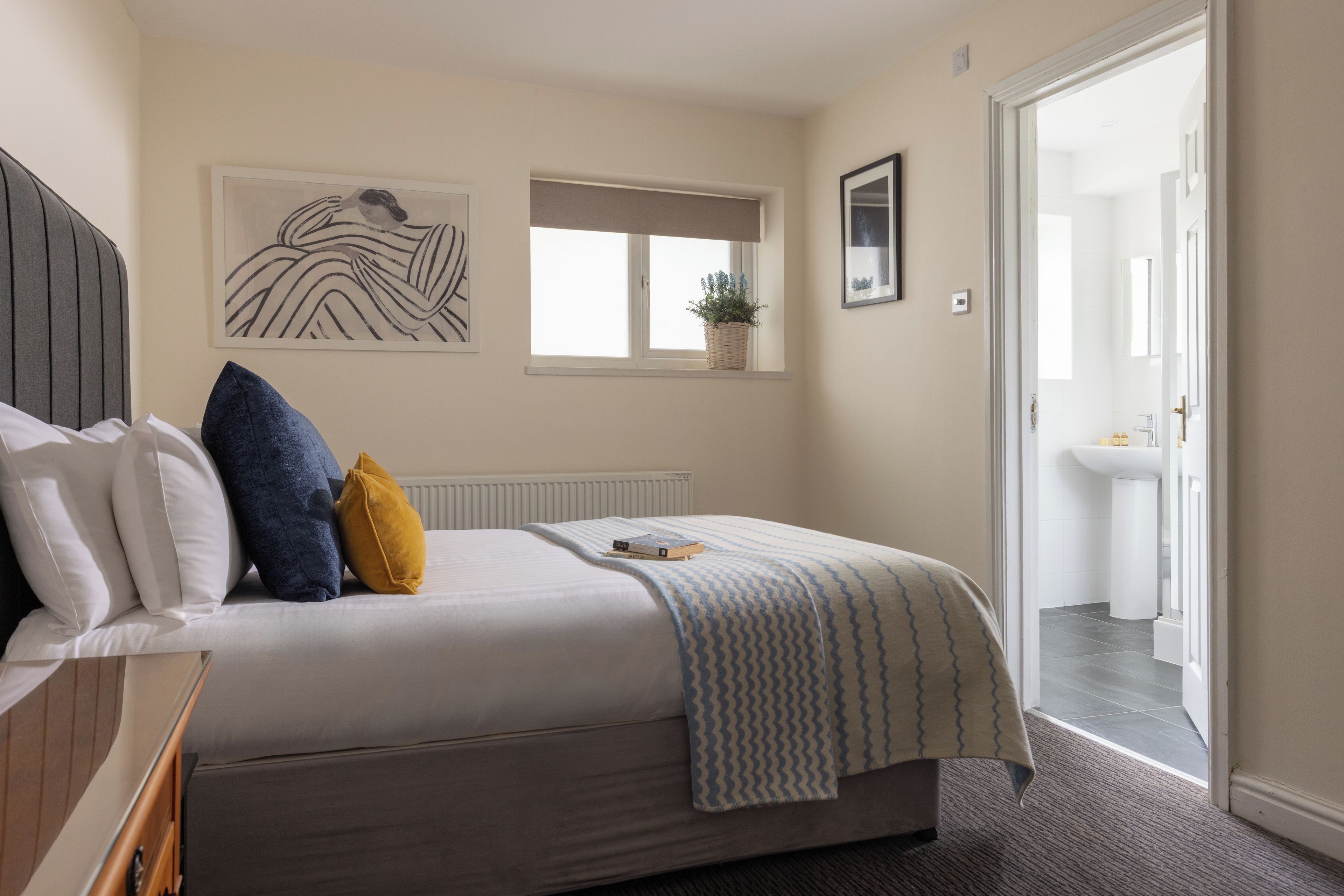 Une chambre à coucher avec un lit, des oreillers, une table de chevet, une fenêtre avec une plante, et une salle de bain visible à travers une porte ouverte.