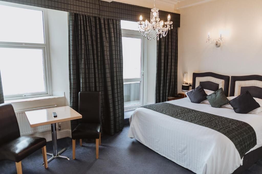 A hotel room with a large bed, a desk, two chairs, a chandelier, and a view of the outdoors.