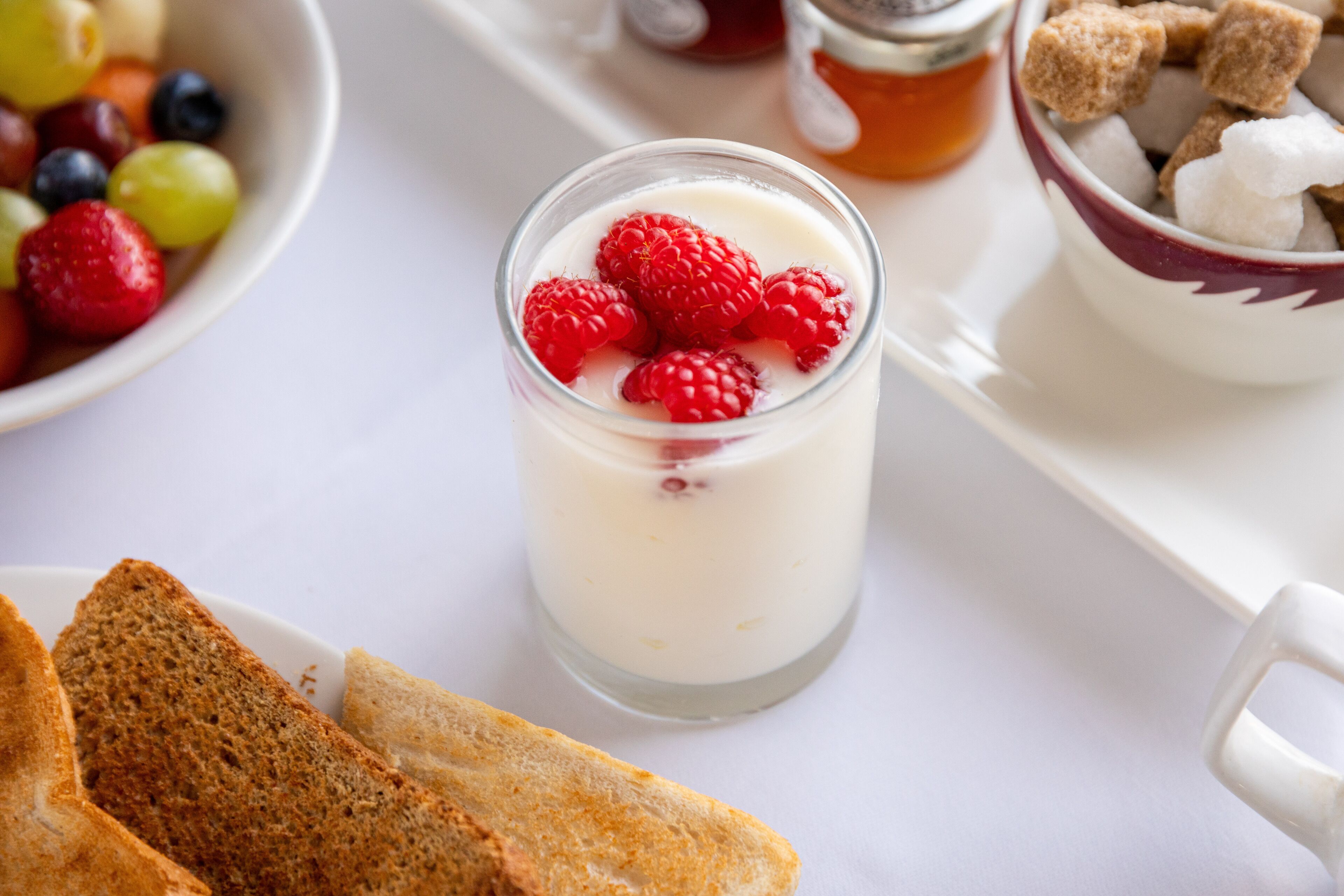 A breakfast setting with a glass of yogurt topped with raspberries, a bowl of mixed berries, and a jar of honey.