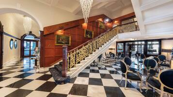 A grand lobby with a black and white checkered floor, a staircase with ornate railings, and a seating area with upholstered chairs.