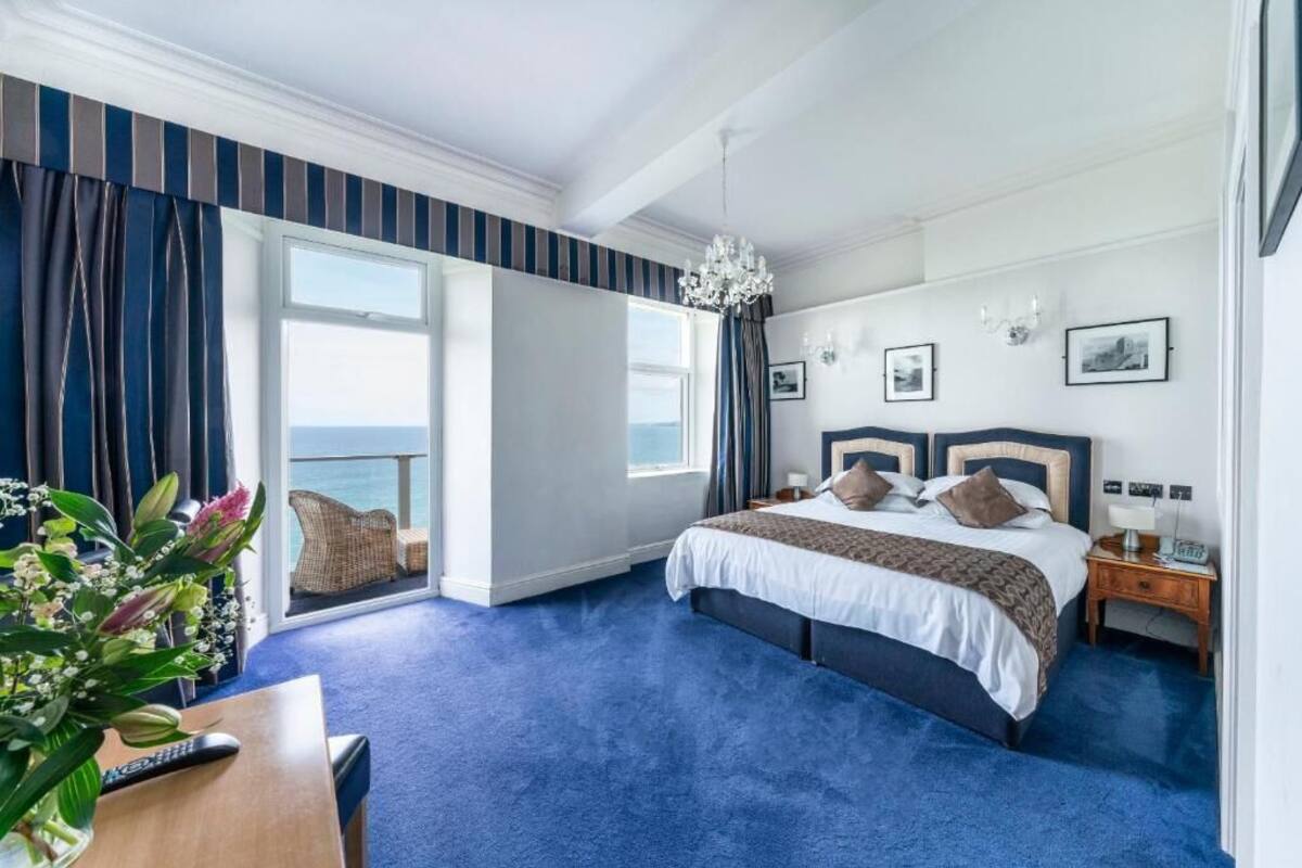 A hotel room with a large bed, a view of the sea, a chandelier, and a wooden nightstand.