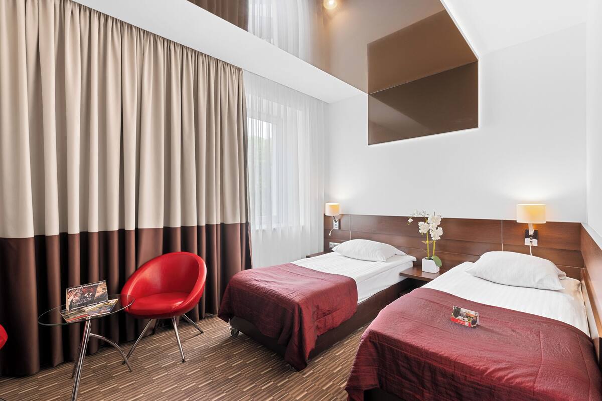 A hotel room with two beds, a red chair, a small table with a magazine, and a window with curtains.