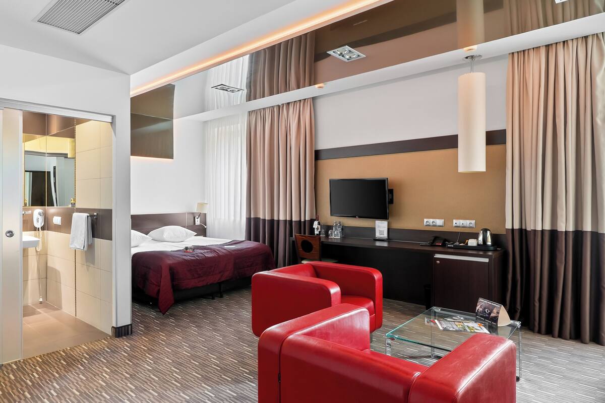 A hotel room with a bed, a desk with a flat-screen TV, a glass coffee table, red leather chairs, a bathroom with a sink and mirror, and a large window with curtains.