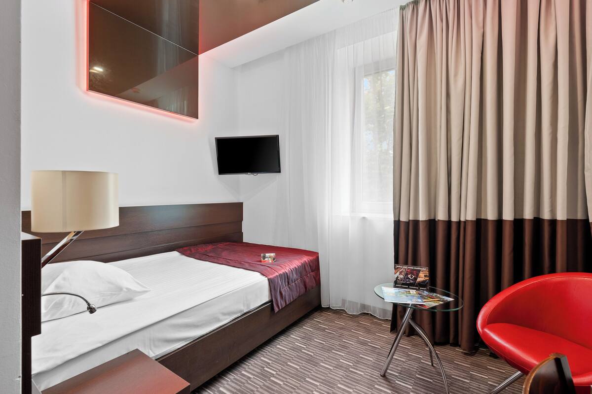 A hotel room with a bed, a TV mounted on the wall, a small table with a magazine, a red chair, and a window with curtains.