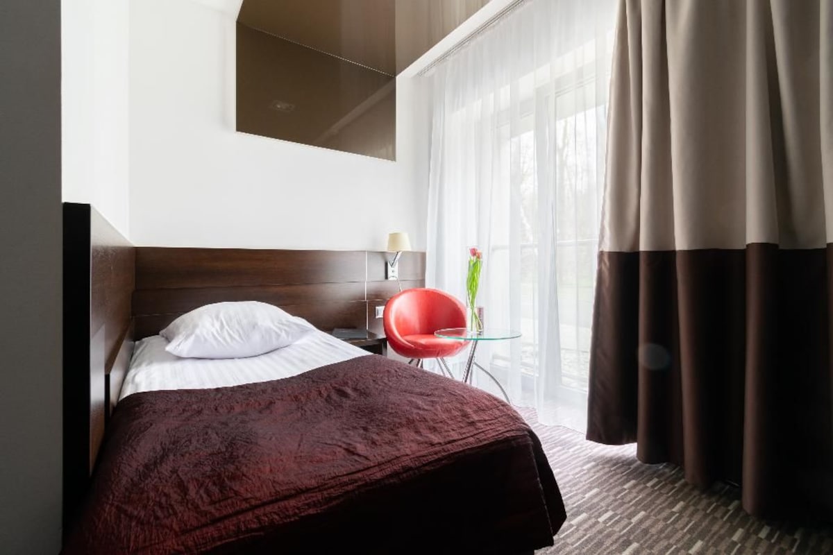 A hotel room with a bed, a red chair, a small table, and a window with curtains.