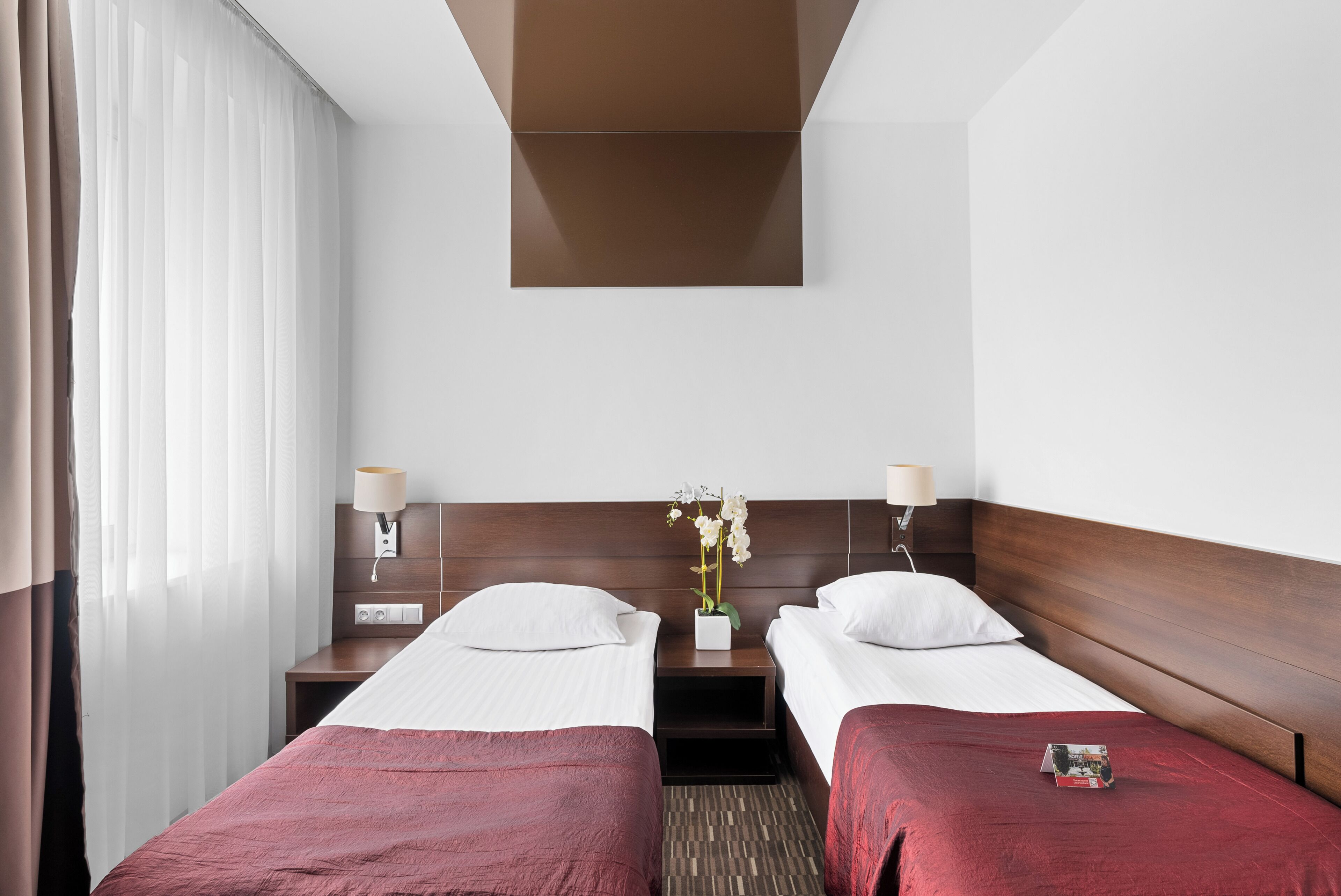 Habitación de hotel con dos camas, una mesita de noche con lámparas, una pequeña mesa con un florero y una ventana con cortinas.