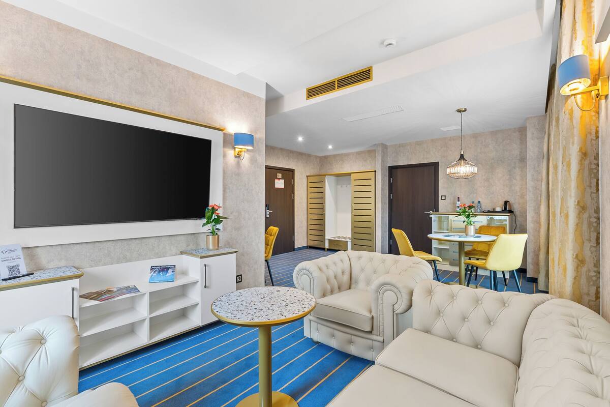 A hotel room with a large flat-screen TV, a seating area with a sofa and armchairs, a small round table, and a kitchenette in the background.