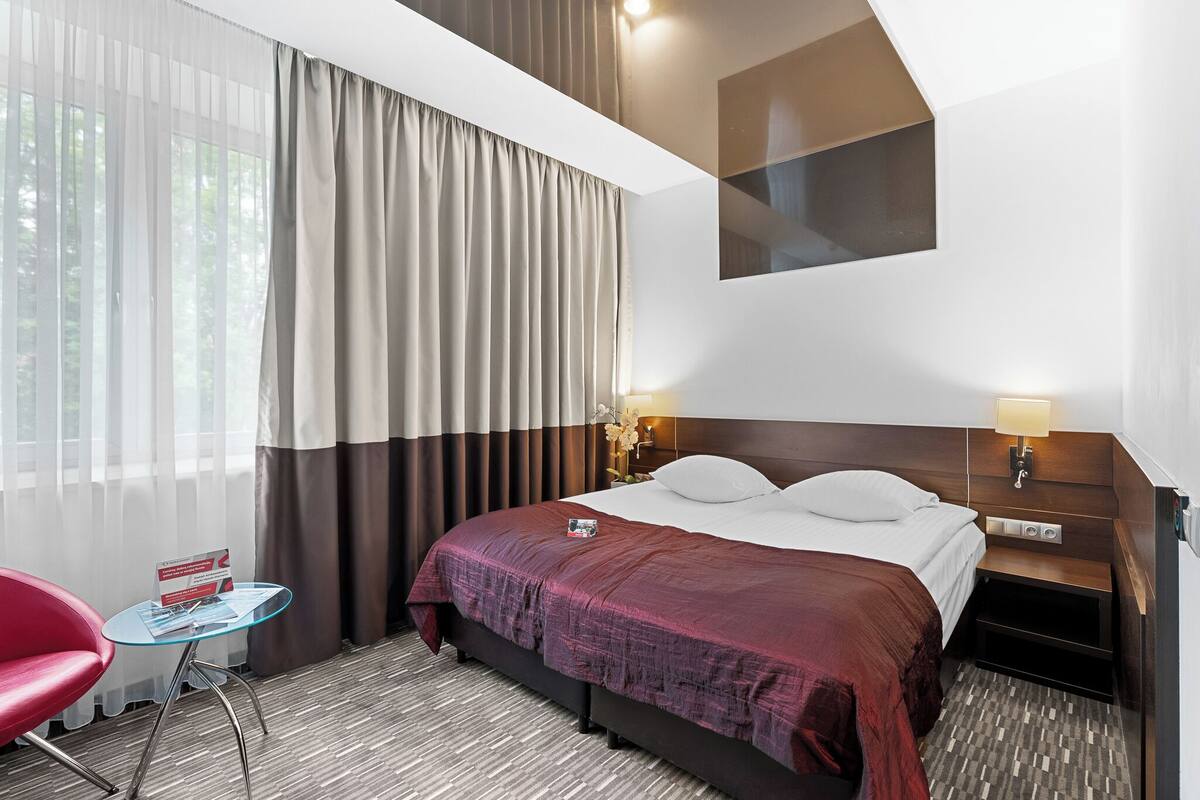 A hotel room with a bed, a red chair, a small table, and a window with curtains.