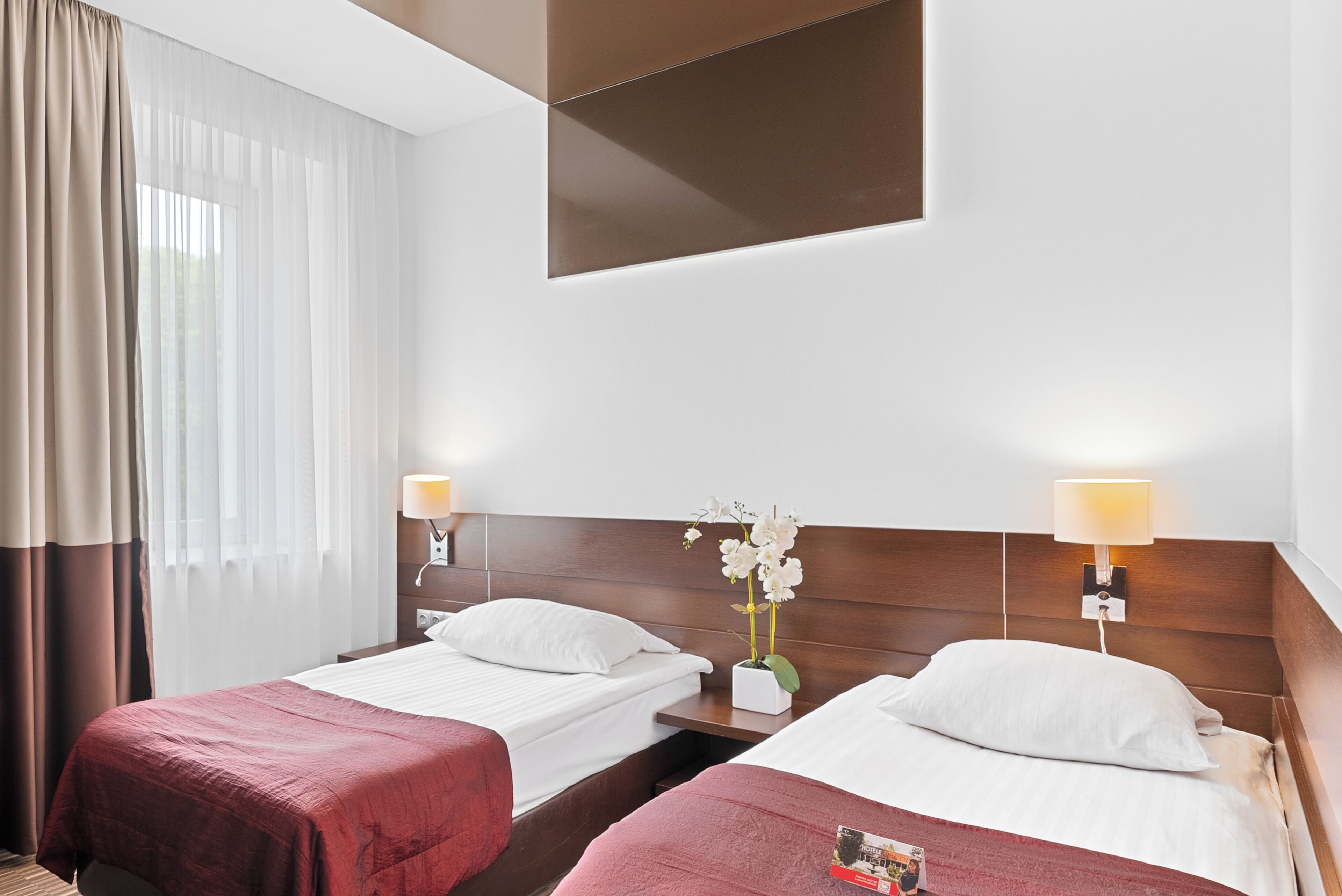 Habitación de hotel con dos camas, cabecera de madera, mesita de noche con lámpara y cortinas traslúcidas.